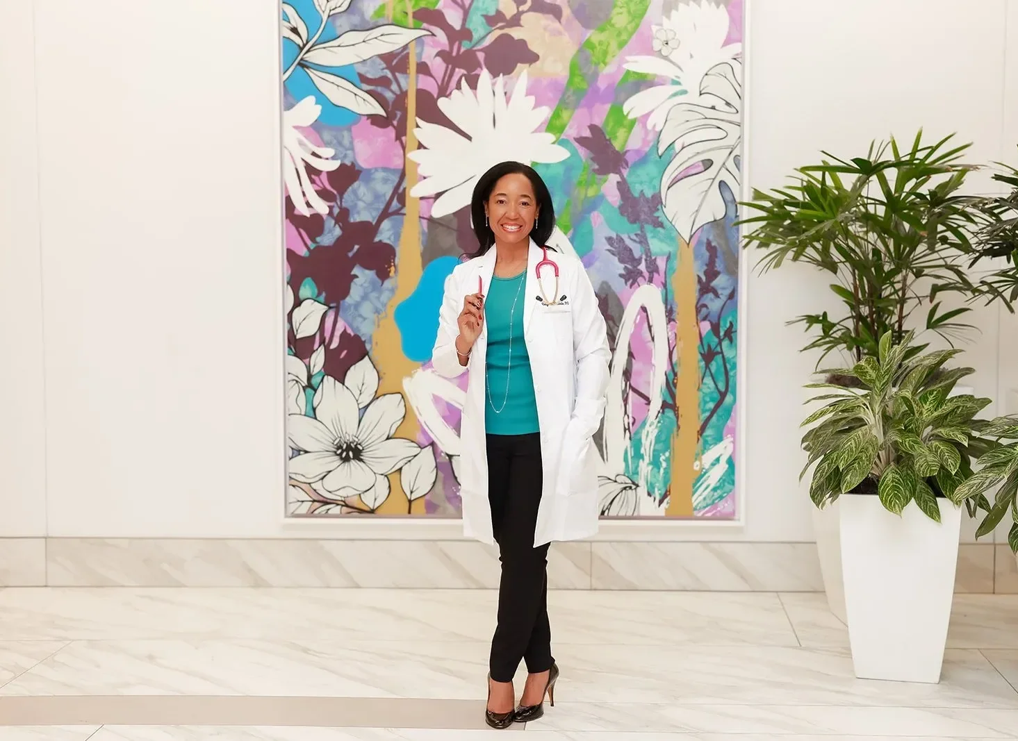 Dr. Betyshia Belardo stands in front of a colorful floral mural, wearing a white coat with a stethoscope around her neck, and black pants.