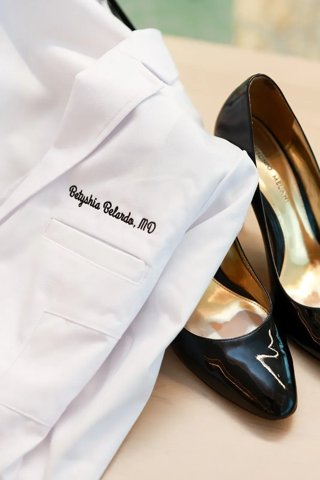 White jacket with embroidered name 'Betyshia Belardo, MD', black patent leather high heel shoes on a table.