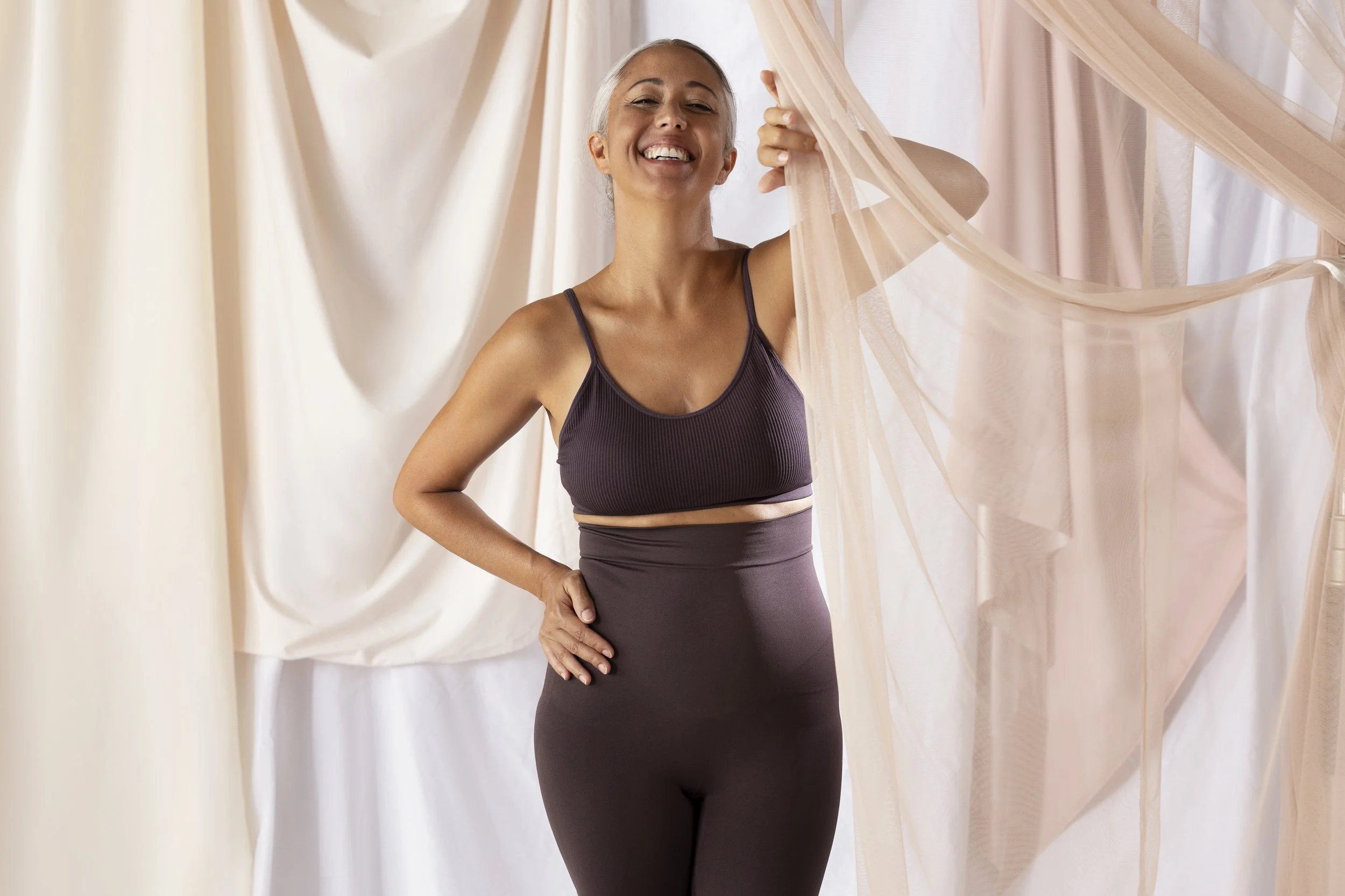 Portrait of a middle-aged woman with gray hair, smiling happily, wearing a dark sports bra and matching leggings, standing among light-colored curtains or fabric drapes.
