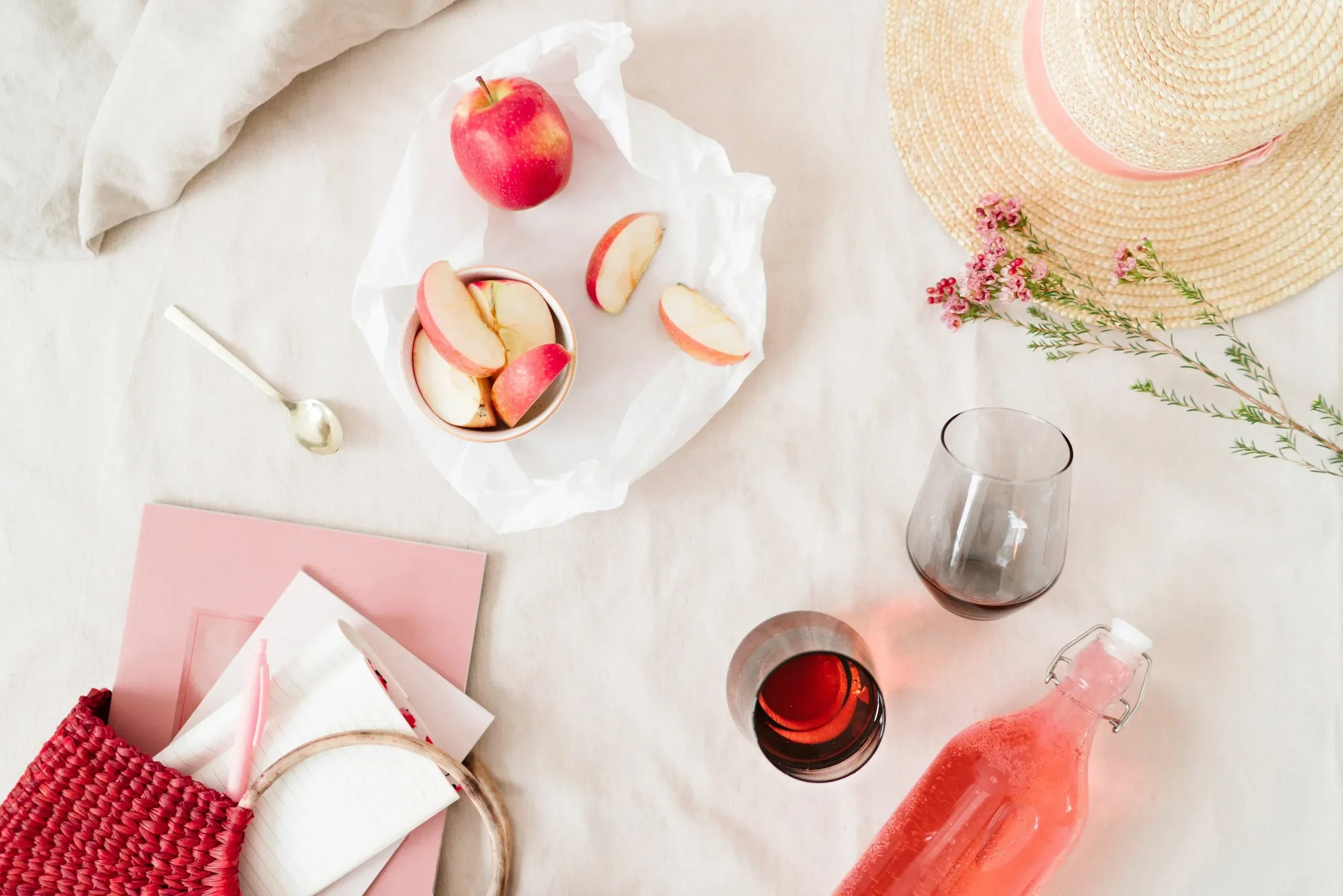A table setting with red apples, a pink straw, pink glasses filled with red drink, a straw sun hat, pink flowers, notebooks, and a red woven bag.