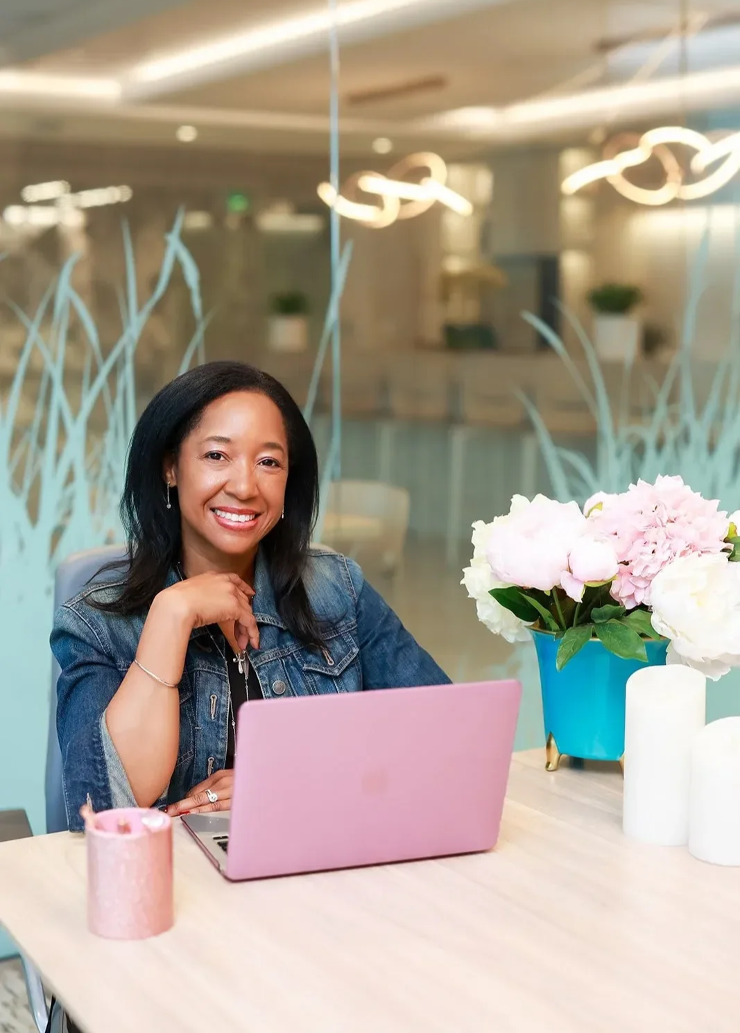 Dr. Betyshia Belardo sitting at a desk with a pink laptop, smiling, with a floral arrangement in a blue vase and pink candles.