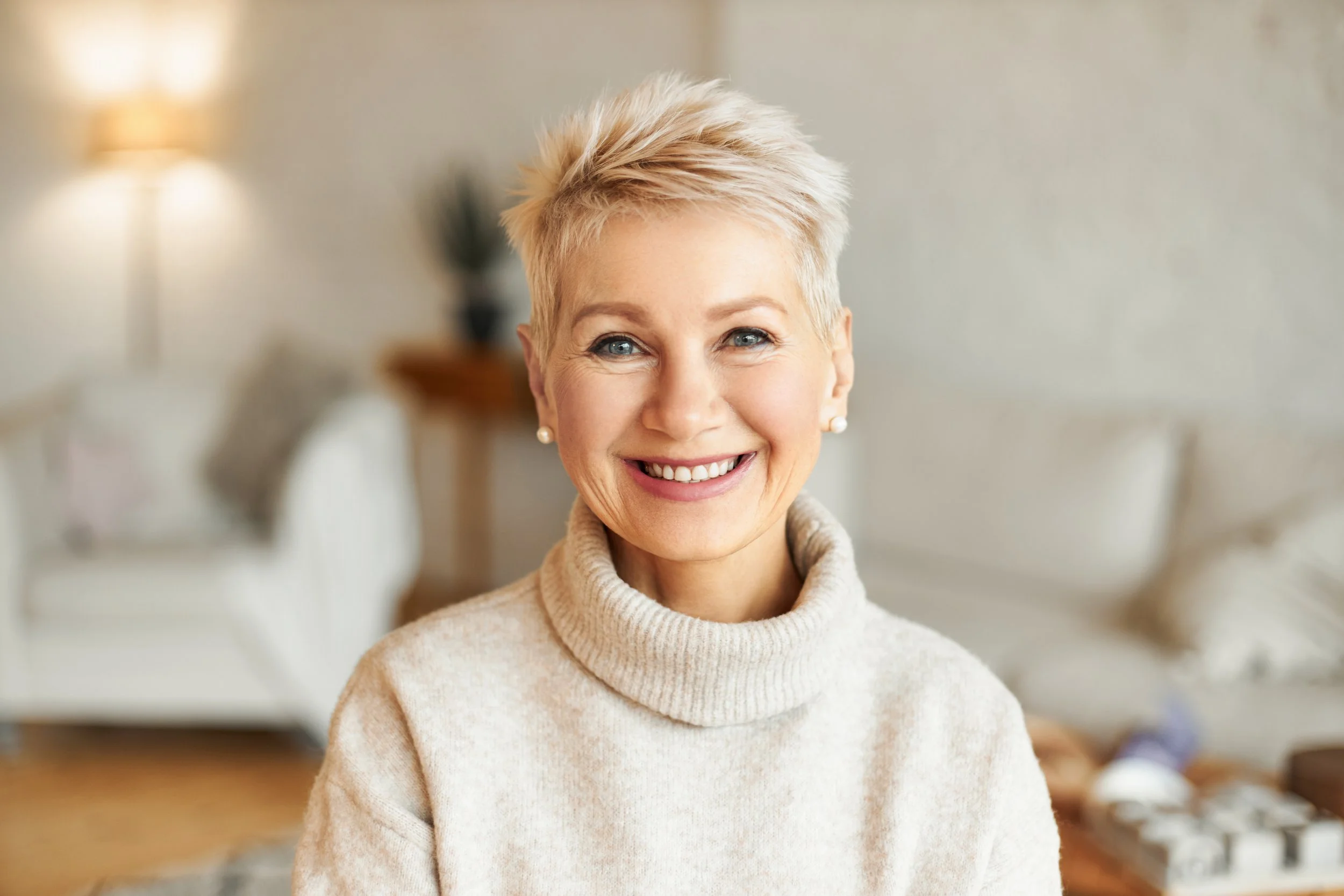Close-up of a smiling woman with short, platinum blonde hair wearing a beige turtleneck sweater.