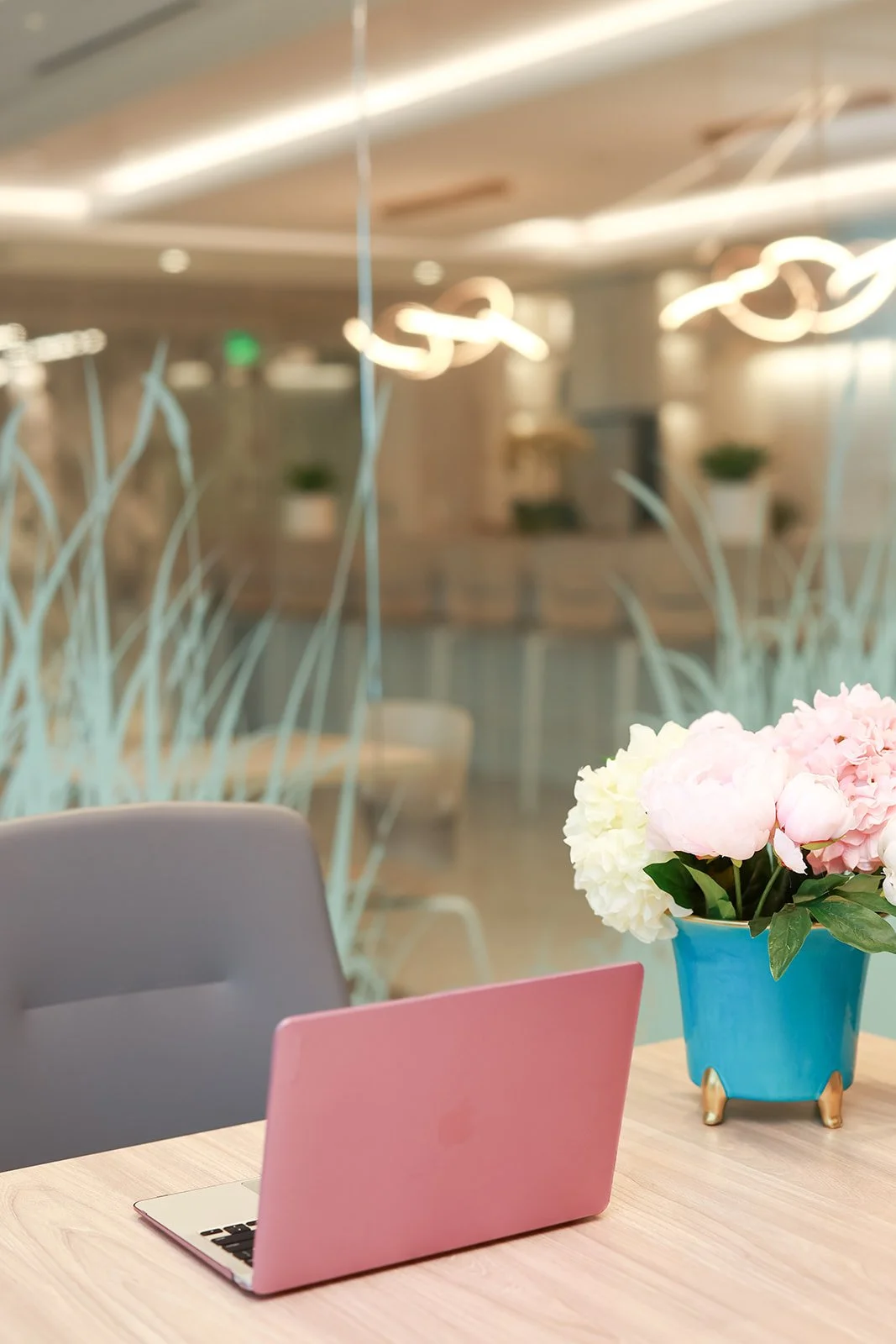 A pink laptop on a desk with a blue pot of pink and white flowers, in a modern office or cafe setting with blurred background.