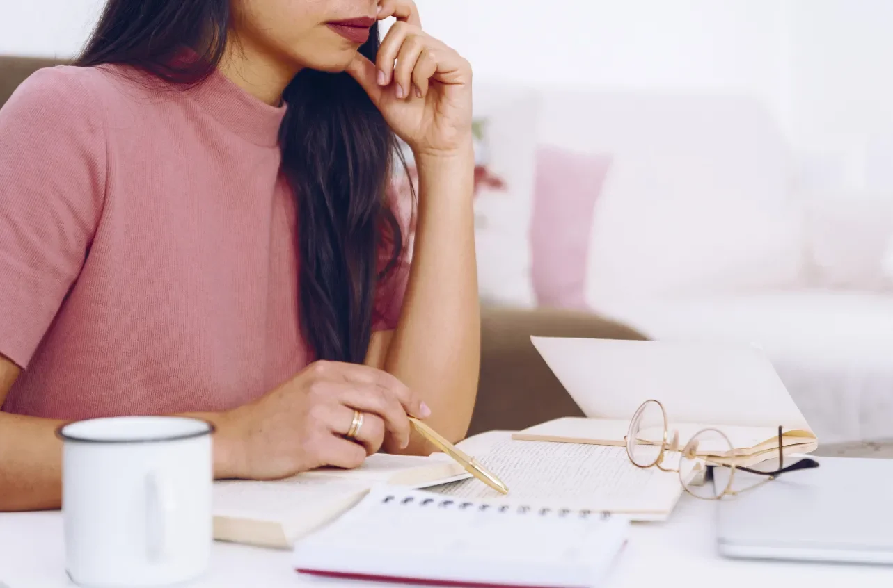 Woman in pink shirt studying at a desk with a mug, open books, glasses, and a laptop.