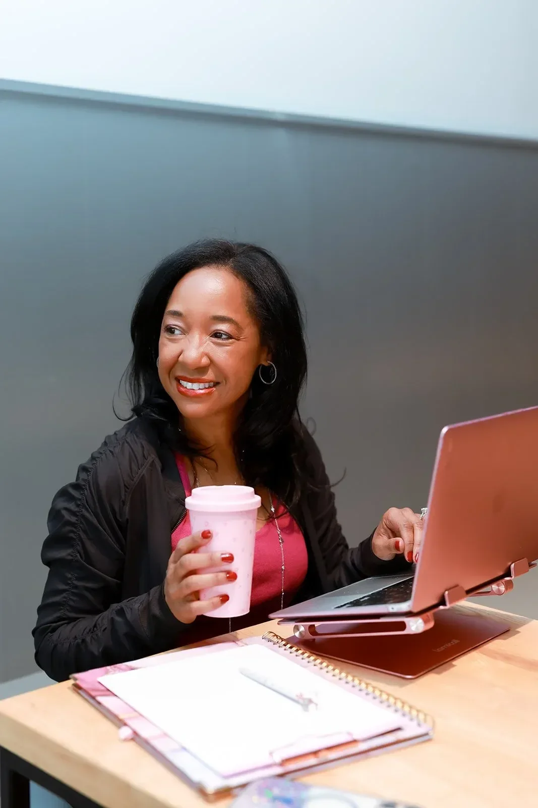 Dr. Betyshia Belardo smiling, seated at a desk with a pink laptop, holding a pink polka-dot tumbler, with notebooks and a pen on the desk.