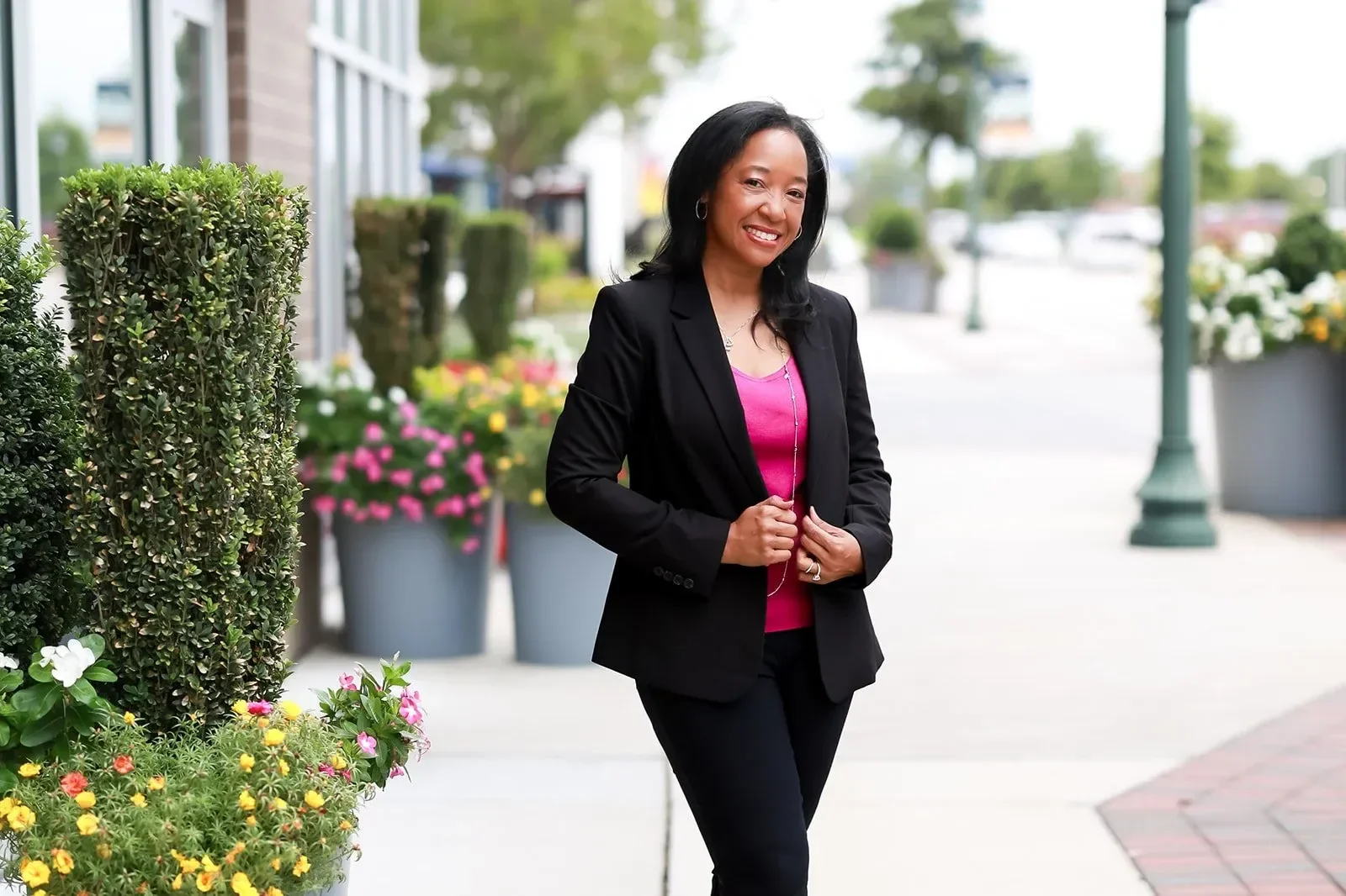 Dr. Betyshia Belardo in a black blazer and pink top walking outdoors on a sidewalk lined with large flower pots and greenery, smiling.