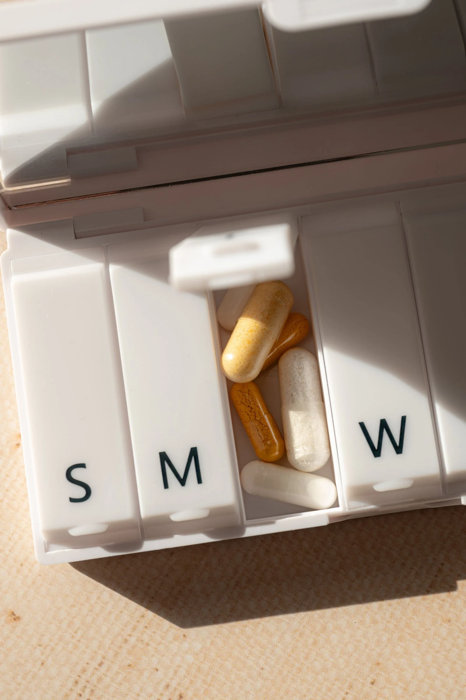 Close-up of a white medication storage box with pills and capsules inside, labeled with letters S, M, and W.