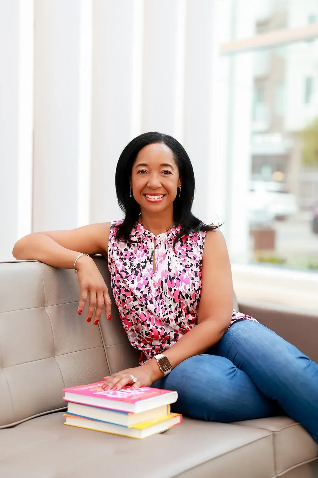 Dr. Betyshia Belardo sitting on a beige sofa, smiling, wearing a sleeveless pink, black, and white patterned top, blue jeans, and a smartwatch, with a stack of books beside her in a bright room with large windows.
