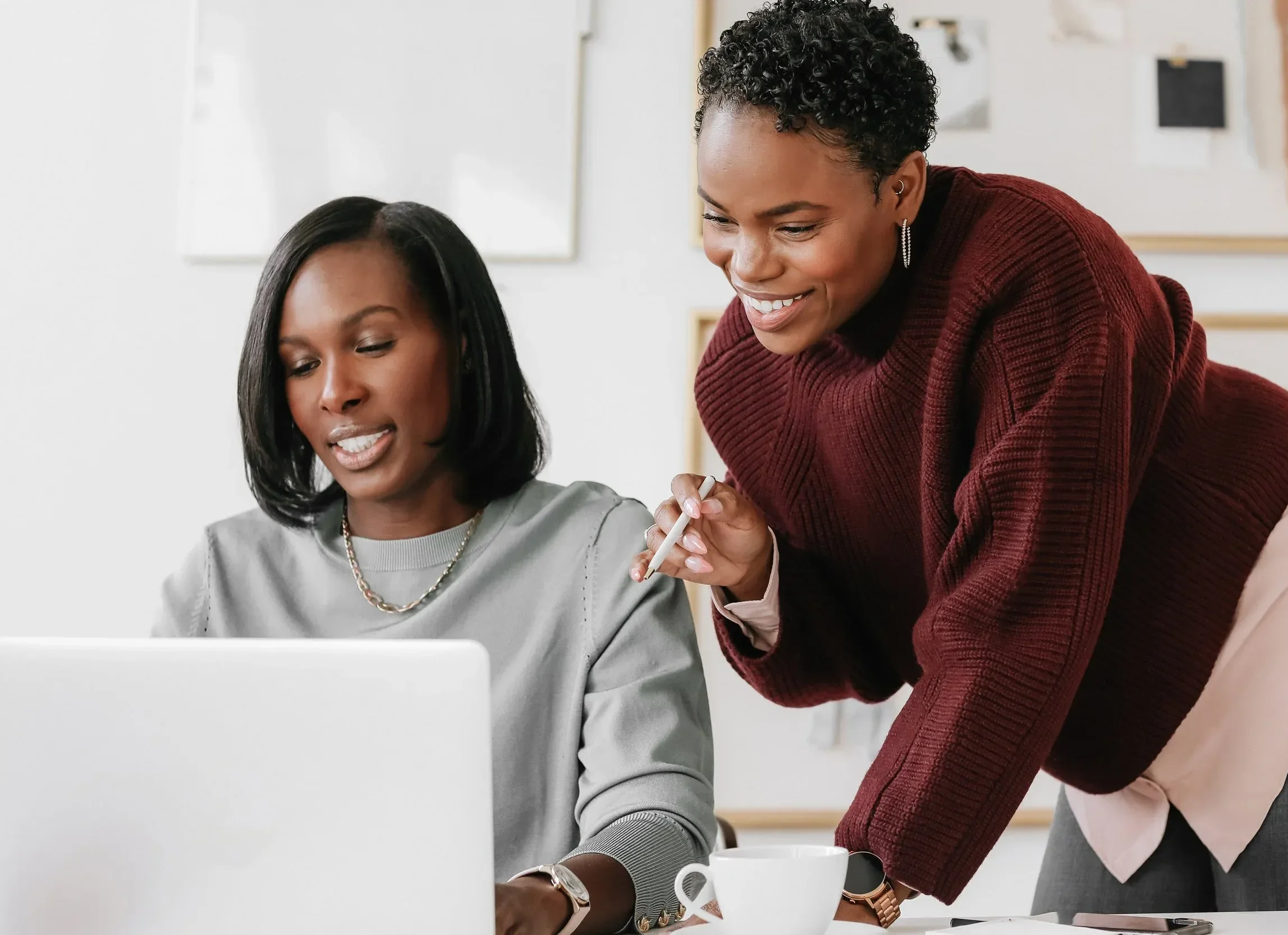 Two women working together on a laptop in an office environment, with one woman standing and pointing at the screen, and the other woman sitting and looking at the computer.