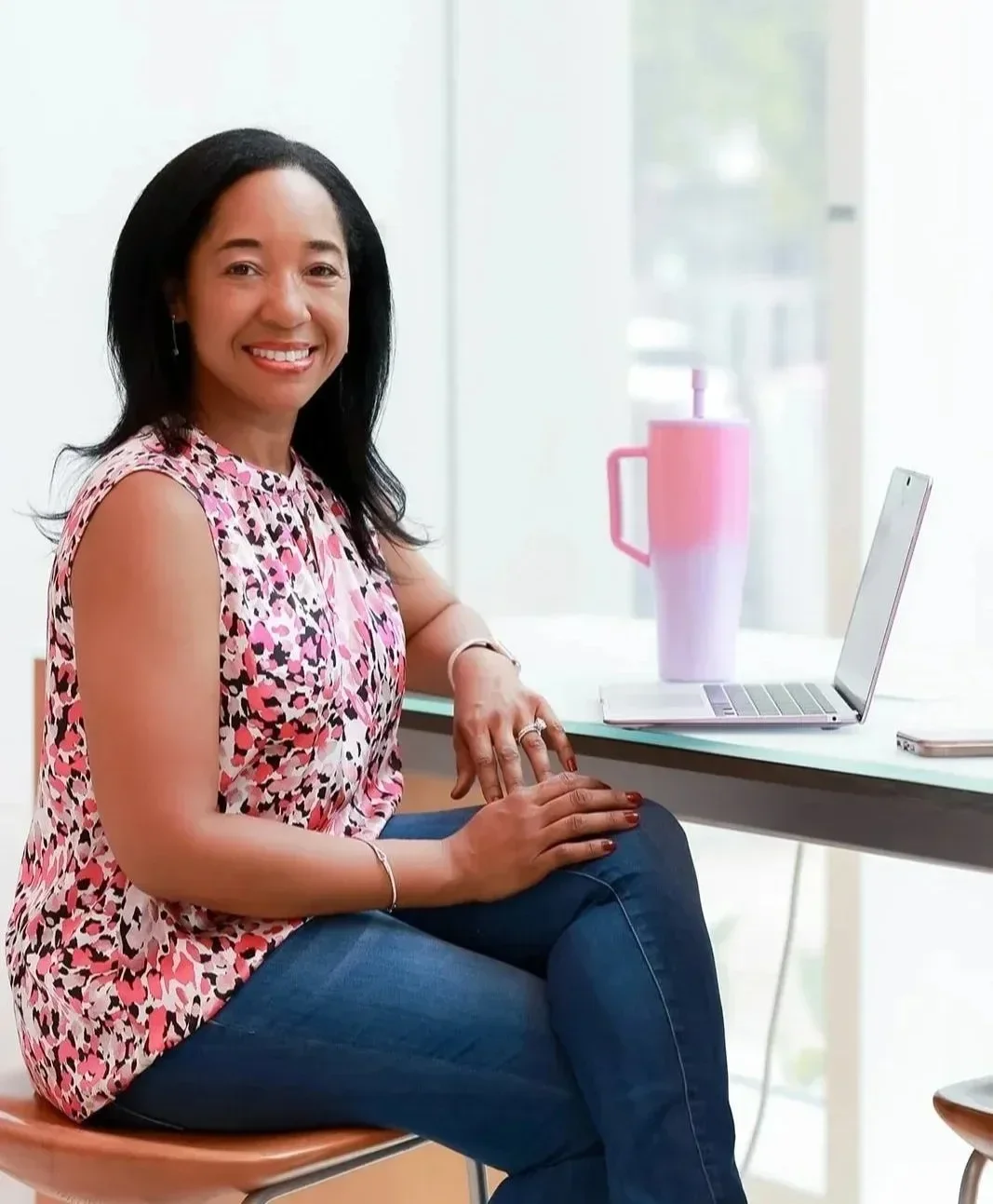 Dr. Betyshia Belardo sitting at a white table with a silver laptop and a pink water bottle, smiling at the camera in a bright room.