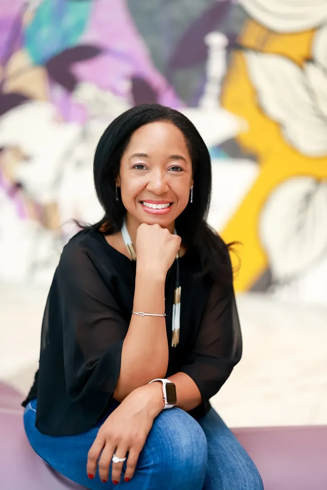 Dr. Betyshia Belardo smiling and sitting with her chin resting on her hand, in front of a colorful mural.