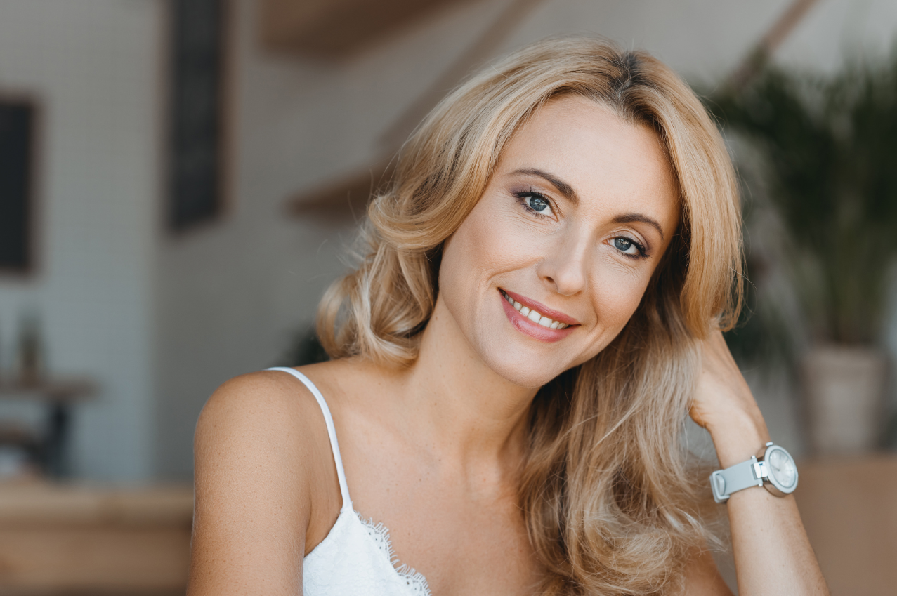 A smiling woman with blonde hair wearing a white tank top, sitting indoors with a blurred background of shelves and plants.