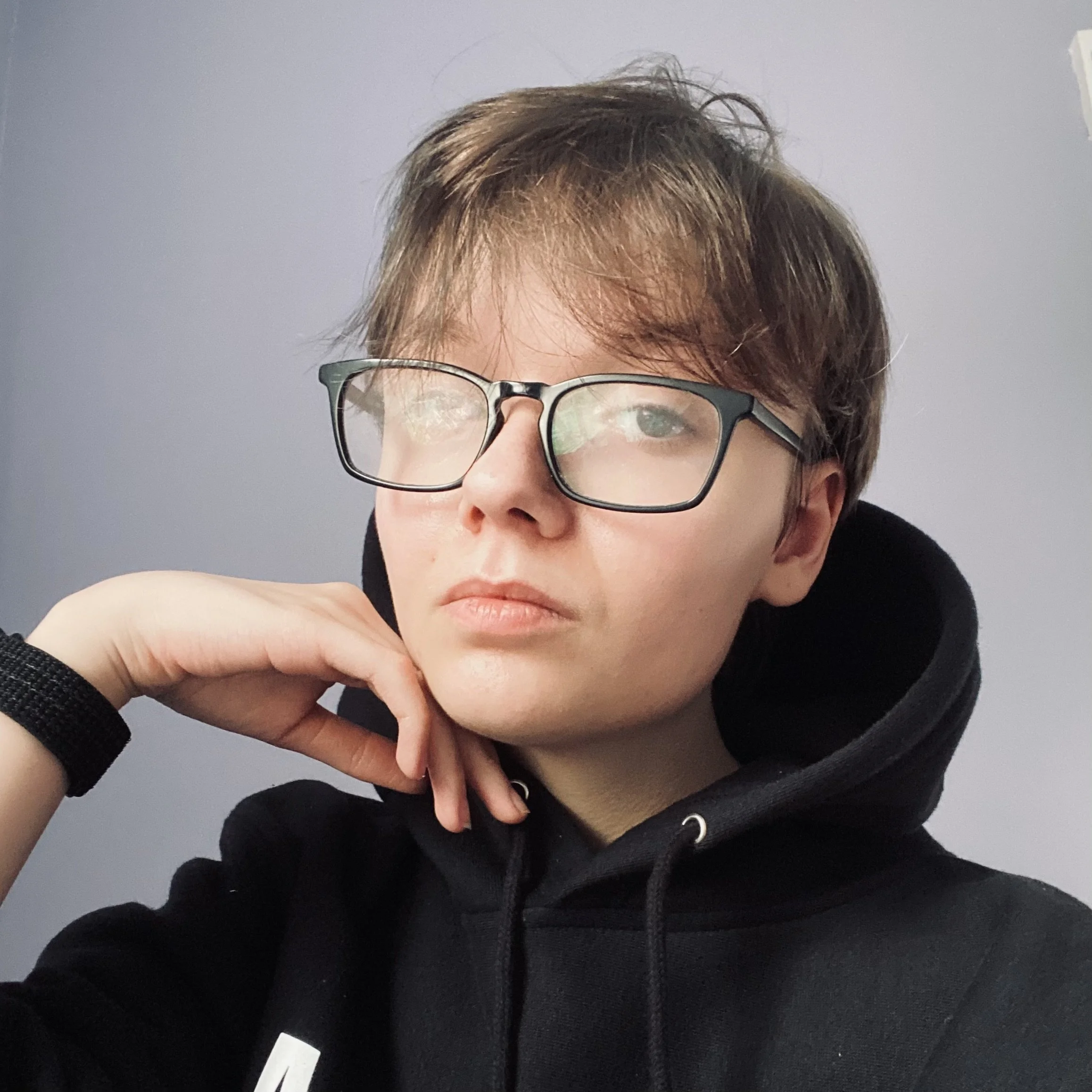 Close-up of a person's face wearing glasses, with short brown hair, resting their chin on their hand, against a gray background.