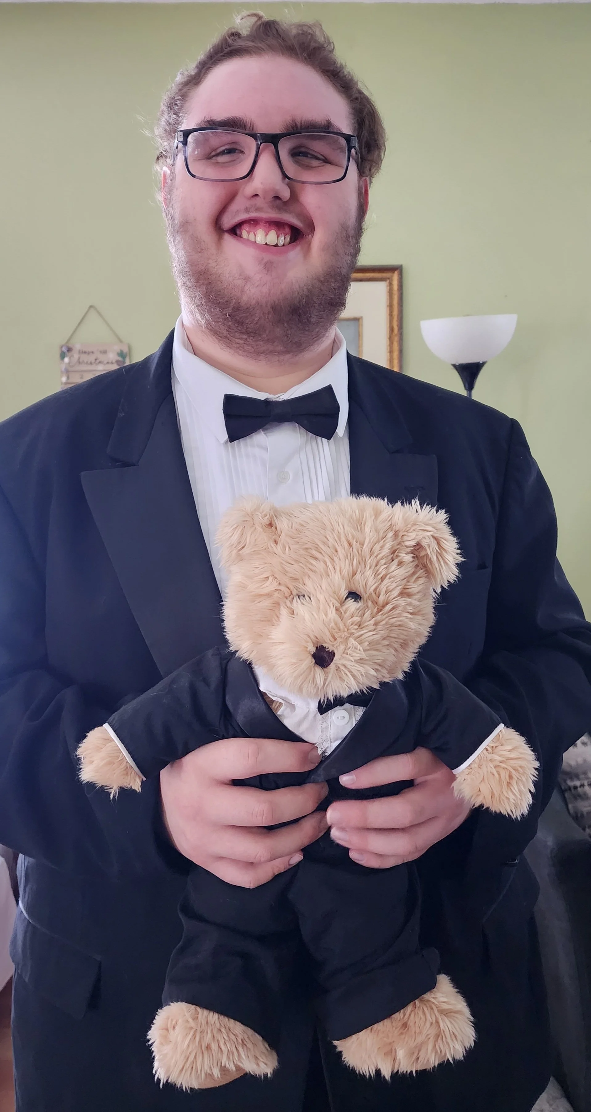 A young man in a tuxedo holding a teddy bear dressed in a tuxedo.