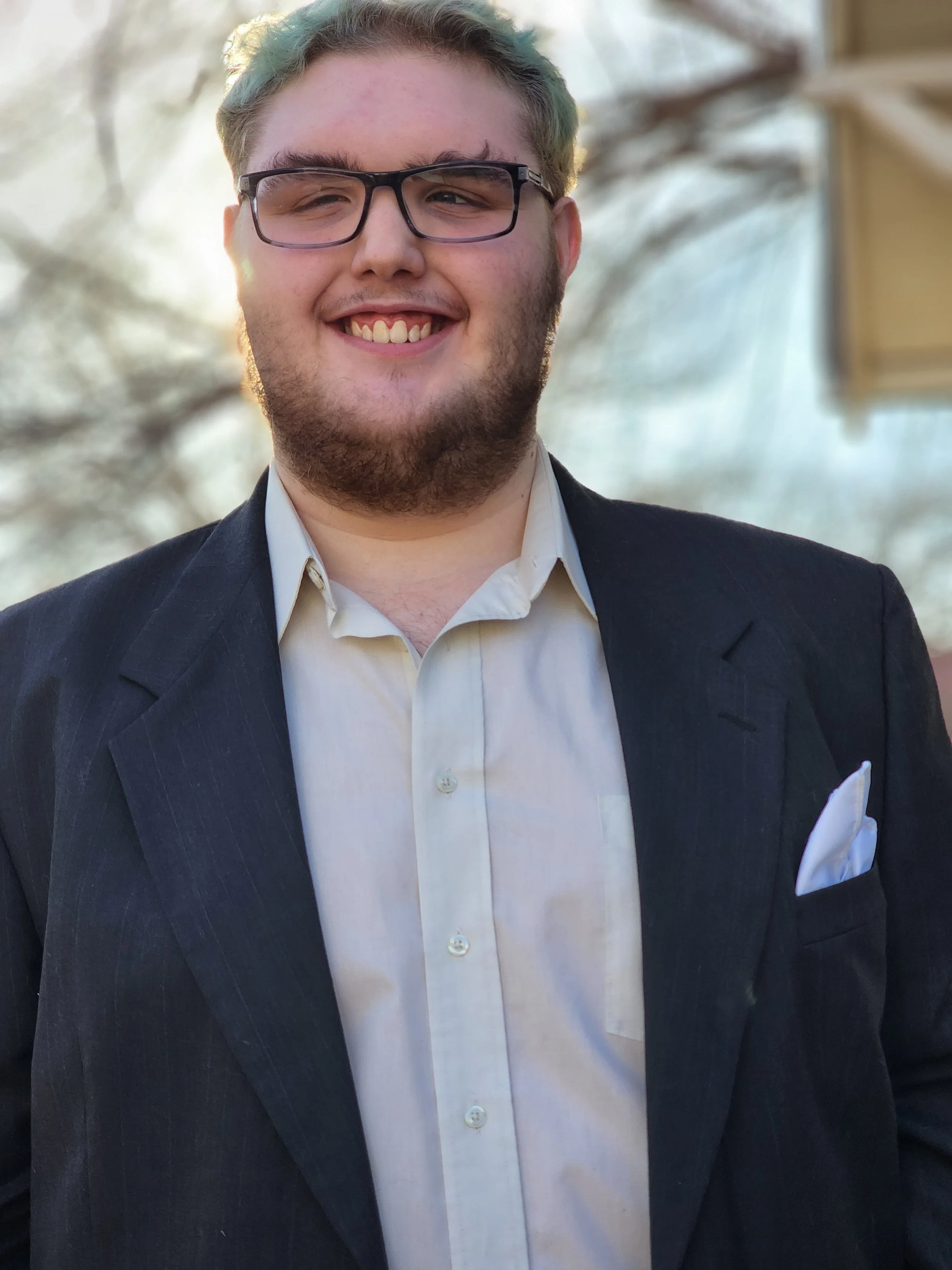 A young man with light skin, glasses, and a beard is smiling broadly, wearing a white shirt and a dark suit jacket with a white pocket square, standing outdoors with blurred trees and a bright sky in the background.