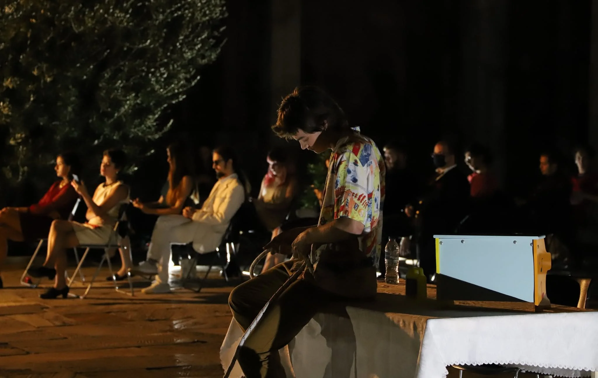 Scene from the operetta ATHANATOS. A group of people sitting outdoors at night, some using phones, with a person in the foreground wearing a colorful shirt and looking down, possibly engaged in a quiet activity.