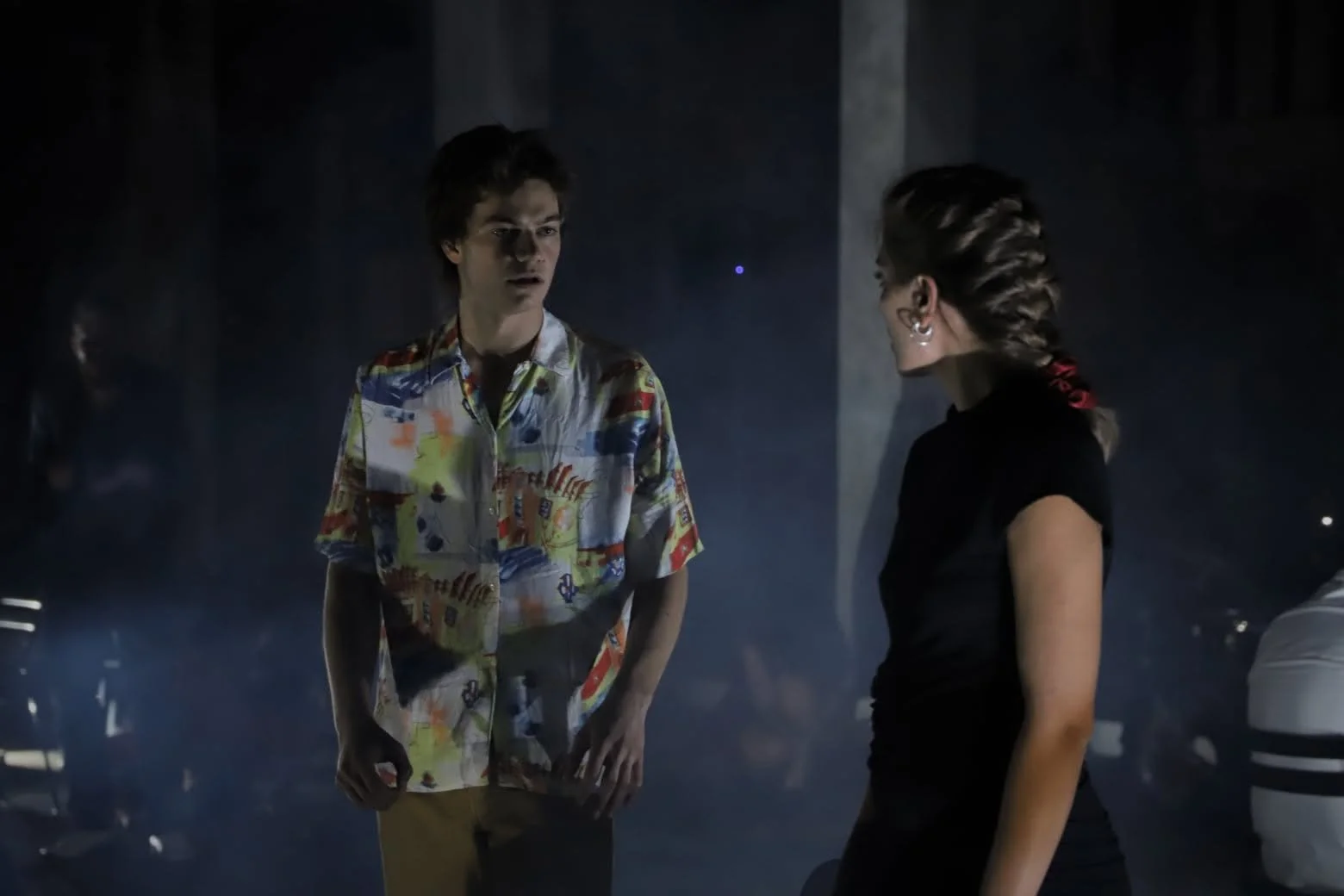Scene from the operetta ATHANATOS. A young man and woman are standing and talking in a dimly lit room with a smoky atmosphere. The man is wearing a colorful patterned shirt, and the woman has her hair in a braid and is wearing a black top.