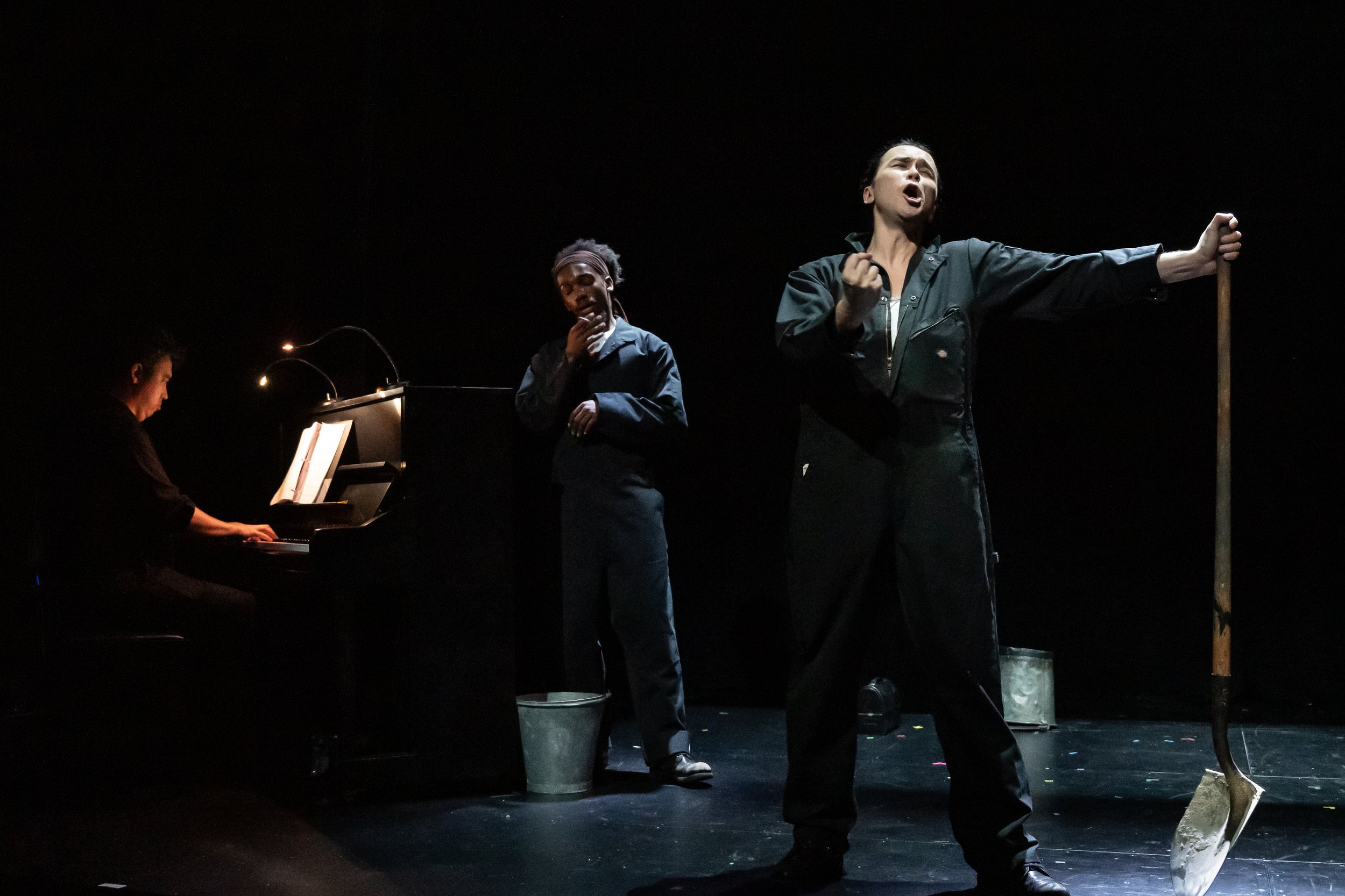 Scene from the opera LOVE, GEORGE. Three performers on stage during a performance, with one playing piano, another singing, and the third holding a prop and singing passionately.