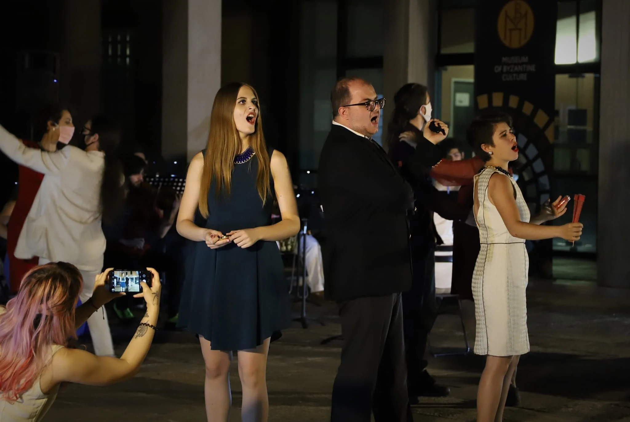 Scene from the operetta ATHANATOS. People singing or performing on stage at an event, some wearing formal attire, with one person recording the performance on a phone.
