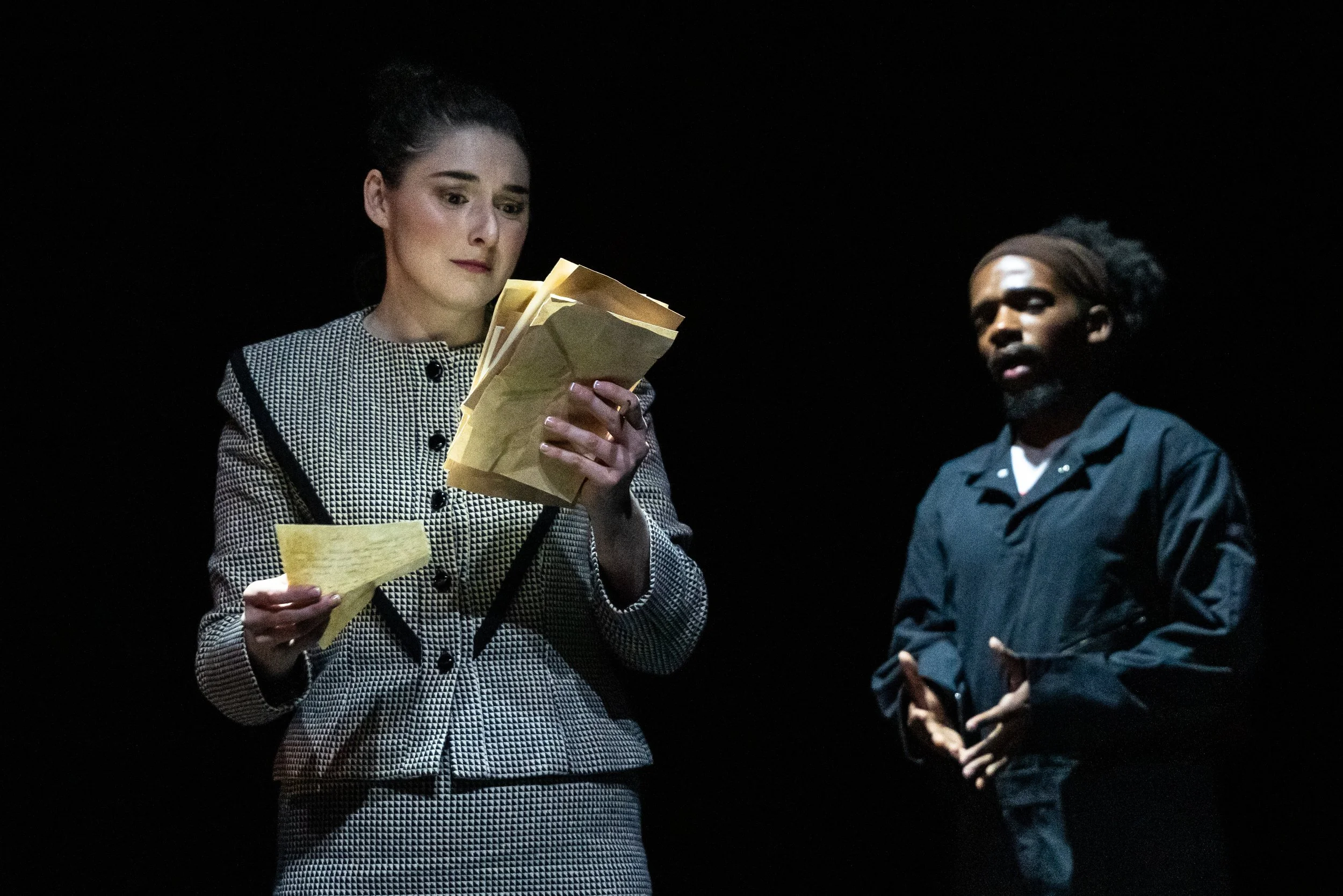Scene from the opera LOVE, GEORGE. A woman with dark hair tied back, dressed in a checkered blazer, is holding papers and reading them, while a man with dark curly hair, glasses, and wearing a dark jacket looks on in a dark setting.