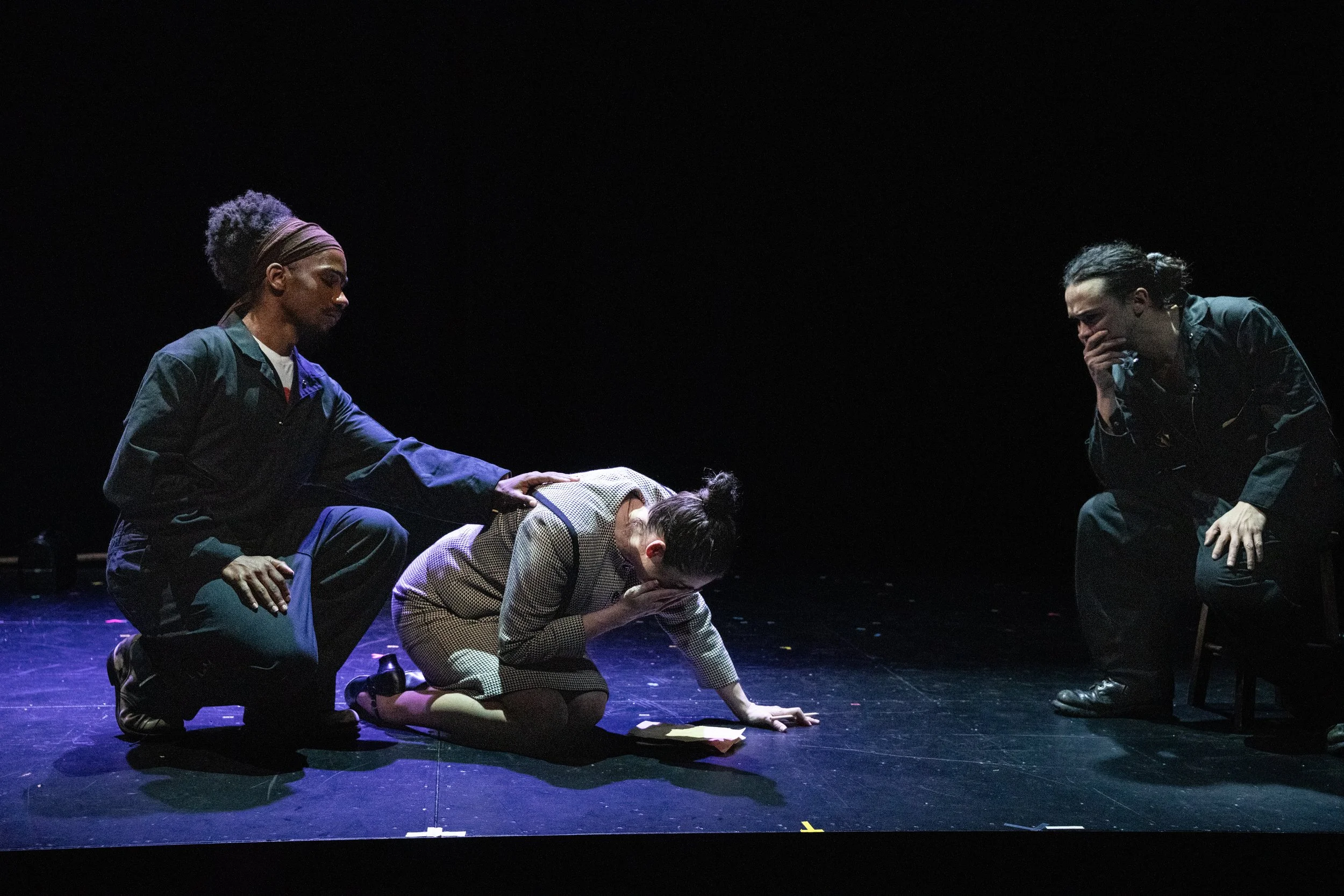 Scene from the opera LOVE, GEORGE.  Four people on stage, one lying on the ground, three kneeling or crouching beside her, in a dark studio setting.