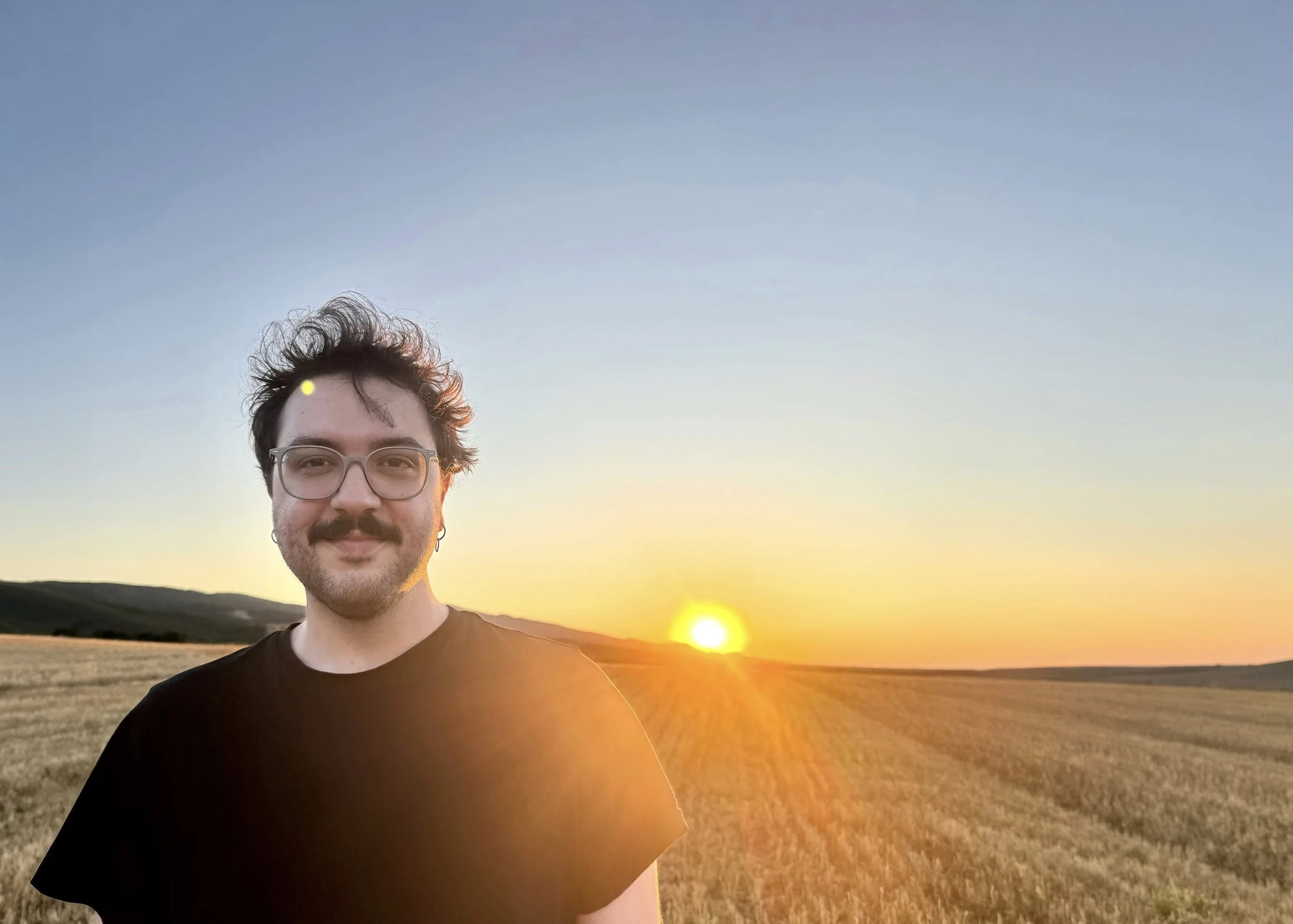 Composer Stefanos Kemanetzis is standing in a field at sunset, with a clear sky overhead and the sun setting on the horizon.