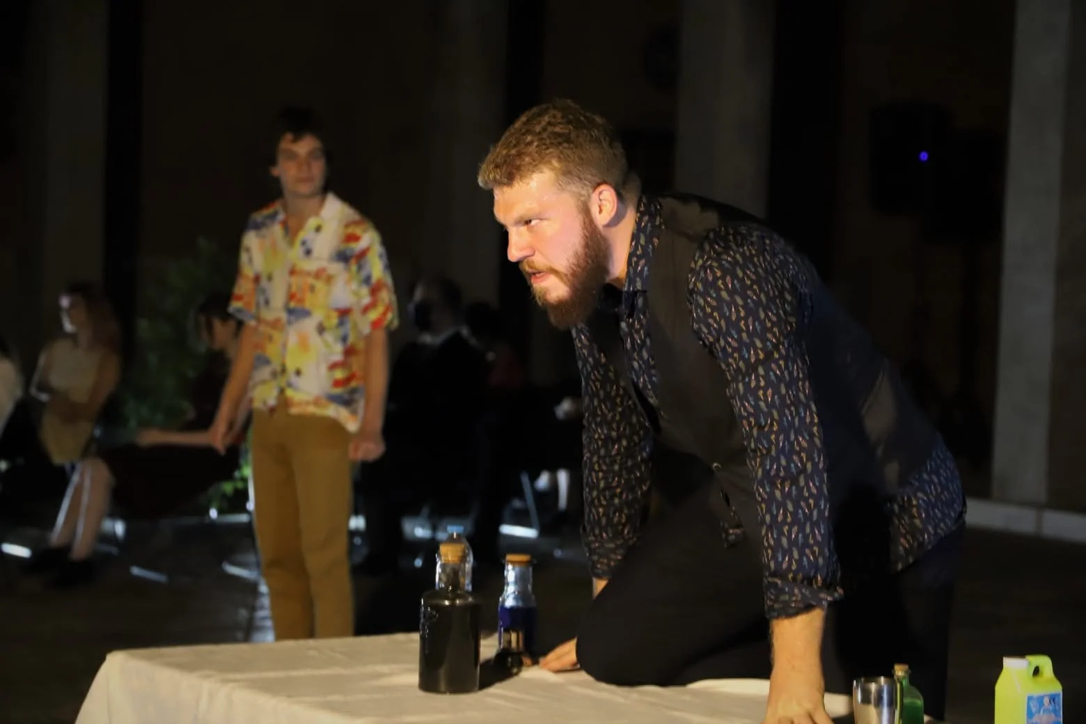 Scene from the operetta ATHANATOS. A man with a beard and patterned shirt bends over a table, with other people in the background at a dimly lit indoor event.