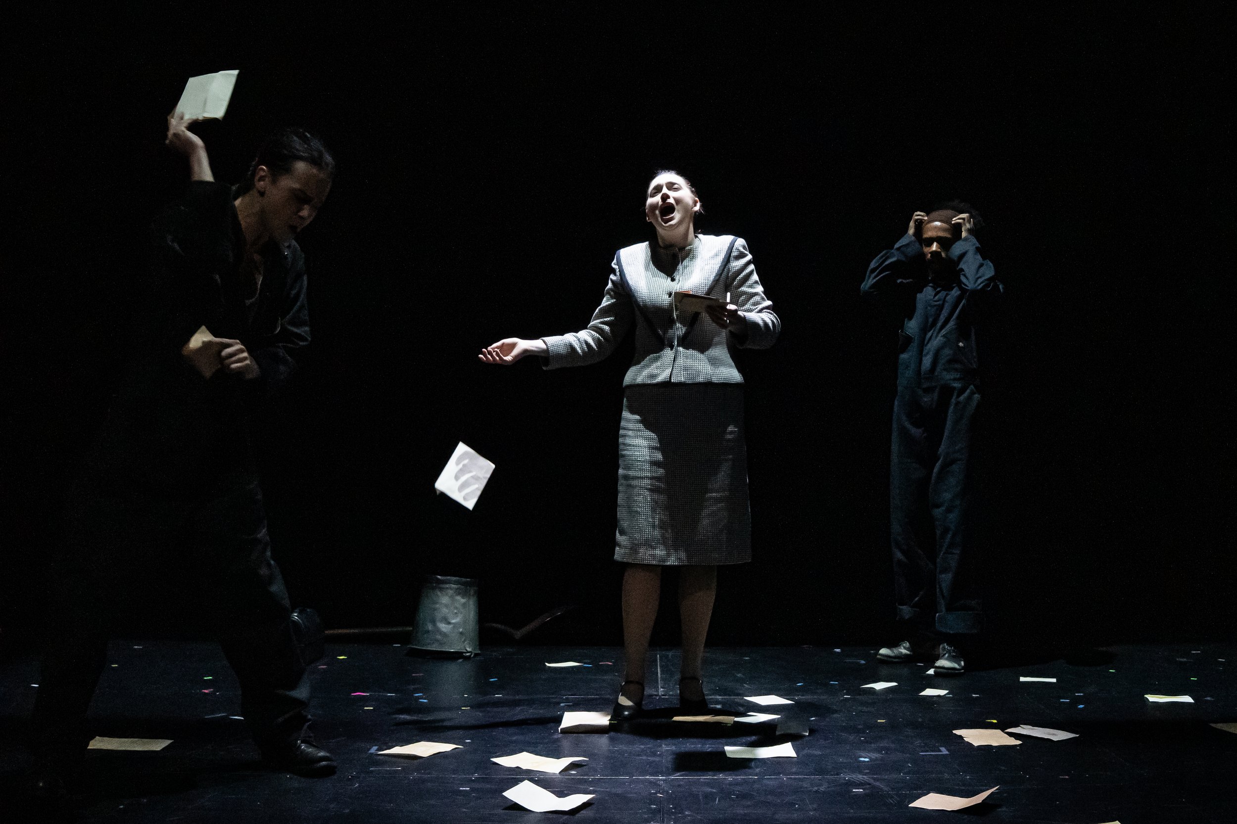 Scene from the opera LOVE, GEORGE. Three people standing on a dark stage, with papers scattered on the floor. The woman in the middle is singing passionately, while the other two are looking away.