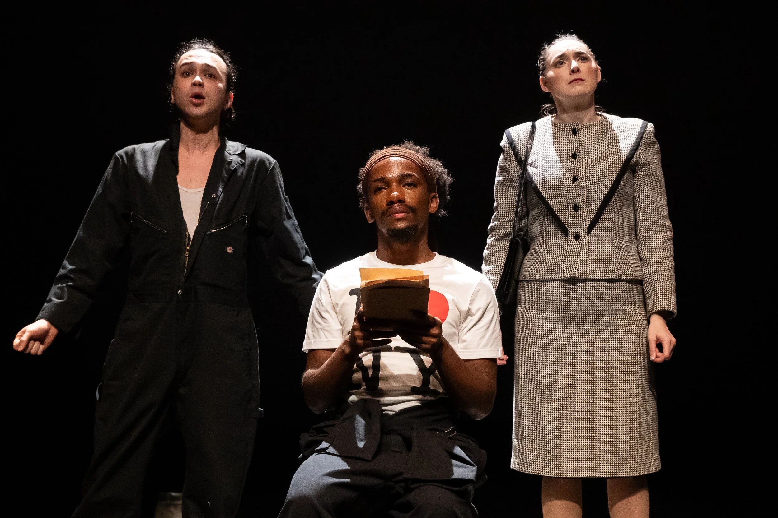 Scene from the opera LOVE, GEORGE. Three singers on stage: one in a black jacket, one in a white I<3NY t-shirt, and one in a plaid suit. The two standing singers appear to be singing, while the seated actor holds a letter.