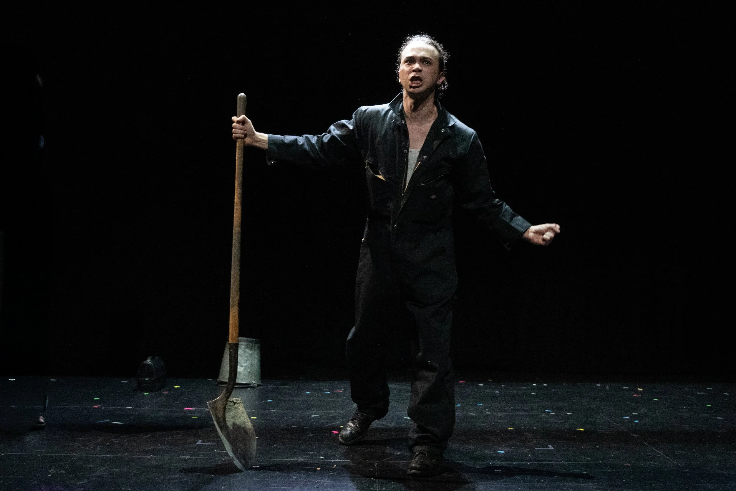 Scene from the opera LOVE, GEORGE. A man in black attire passionately performs on stage, holding a broom upside down with both hands, set against a dark background with colorful confetti on the floor.
