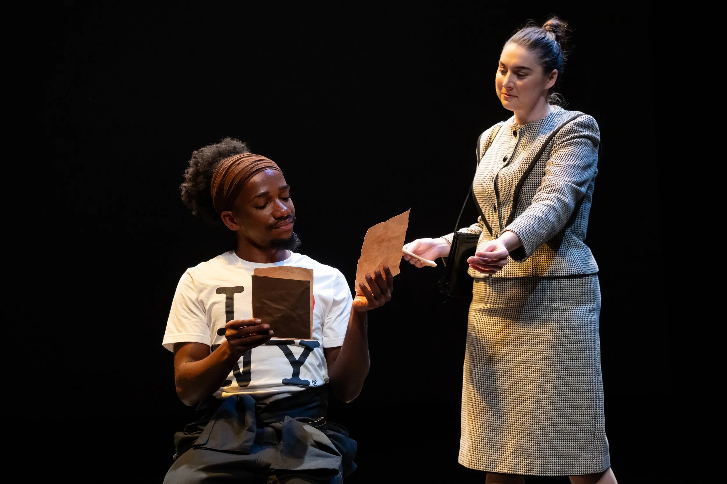 Scene from the opera LOVE, GEORGE. A man sitting on stage receives a paper from a standing woman dressed in a checked suit. The background is black.