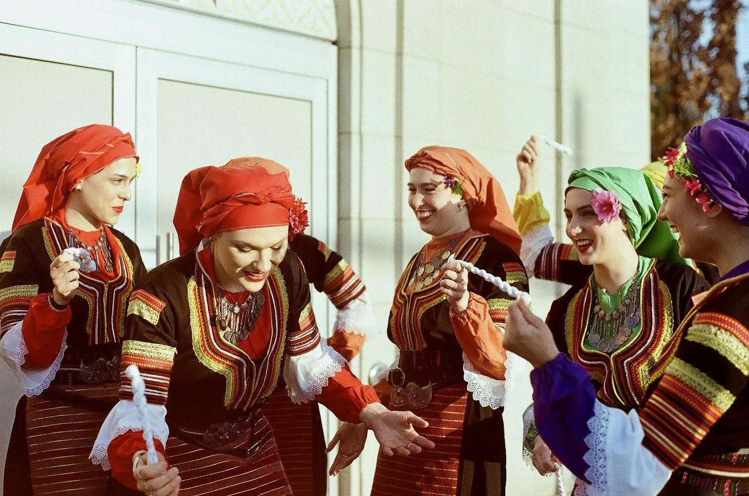 A group of women dressed in traditional folk costumes in black, red, green, purple, and yellow, with colorful headscarves and jewelry, celebrating outdoors and dancing with long white ropes.