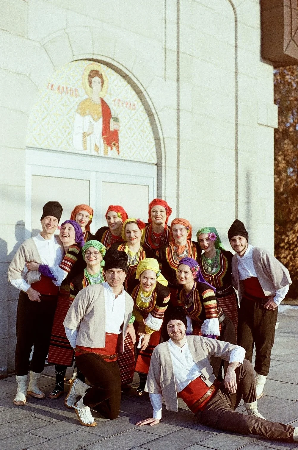 A group of people dressed in traditional folk costumes, smiling and posing in front of a church with an icon above the door.