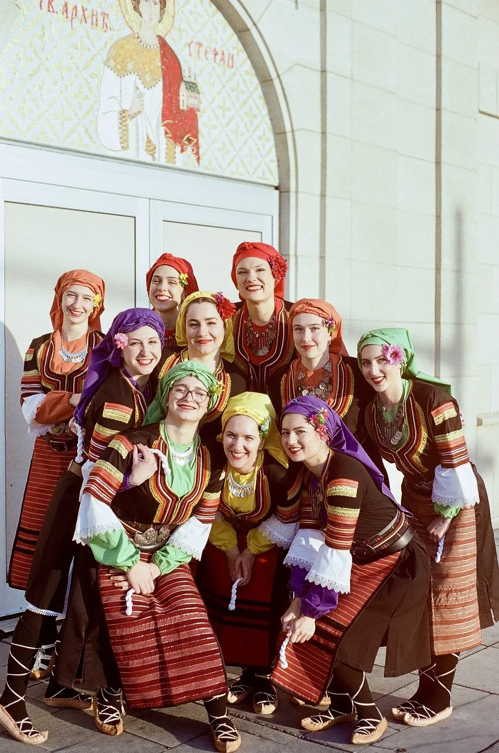 A group of women dressed in traditional colorful folk costumes, standing outside a building with an arched entrance. They are smiling and posing for the photo.