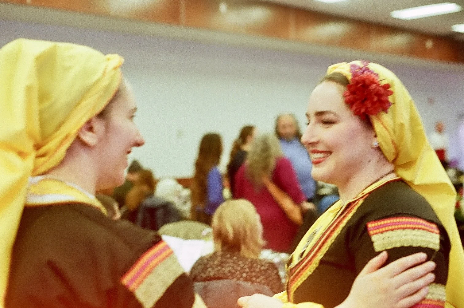 Two women wearing traditional headscarves and costumes are smiling and interacting at a social gathering.