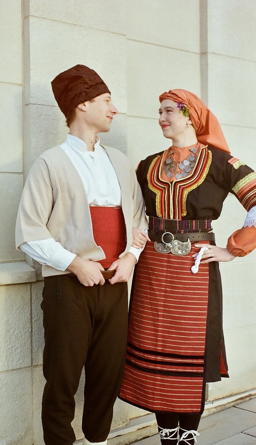 Two people wearing traditional Balkan folk costumes, a young man on the left and a woman on the right, standing outside near a stone wall, looking at each other.