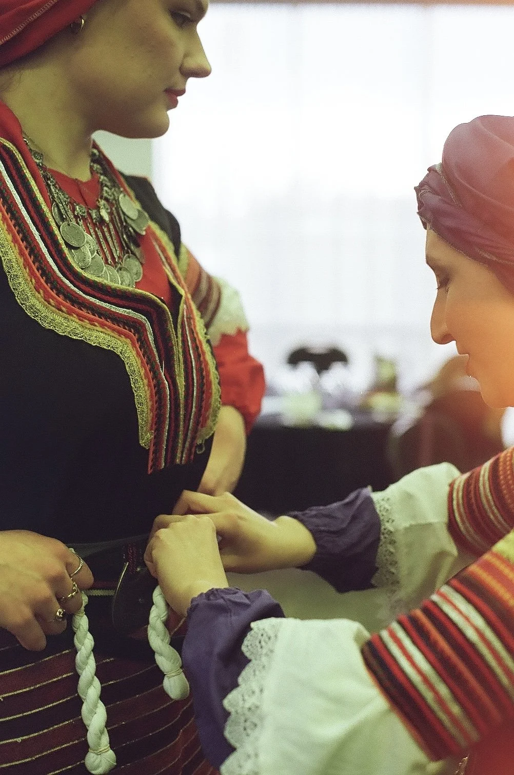 Two women dressed in traditional indigenous attire, with one woman kneeling and tying a braided white rope around the waist of the other woman.
