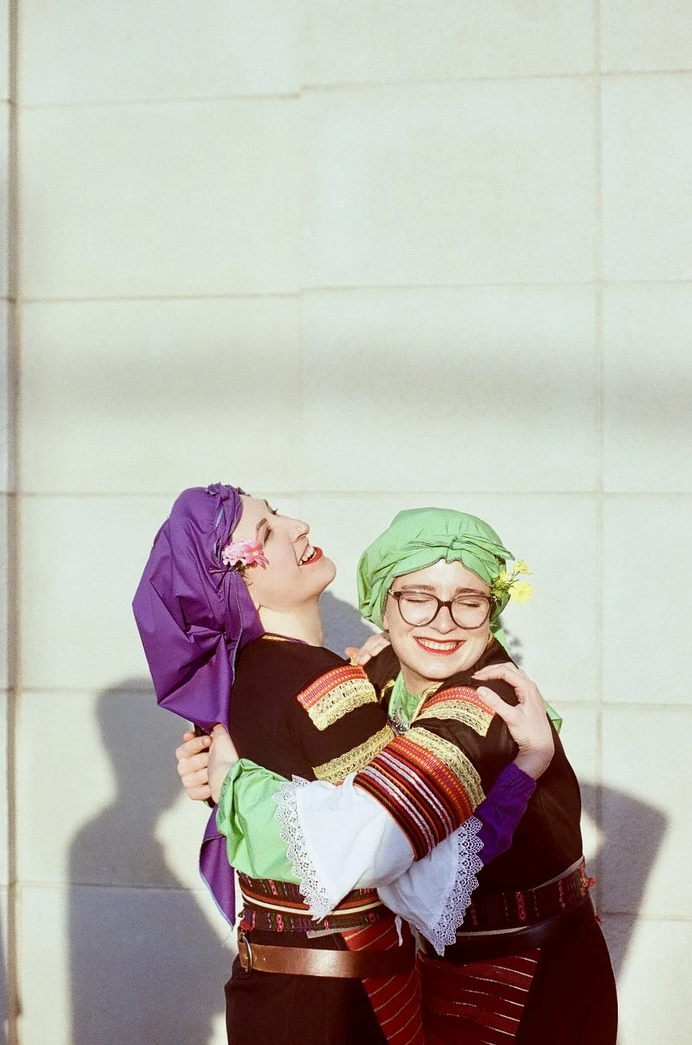 Two women in traditional folk costumes hugging and smiling, each wearing colorful headscarves and glasses, against a plain light-colored wall