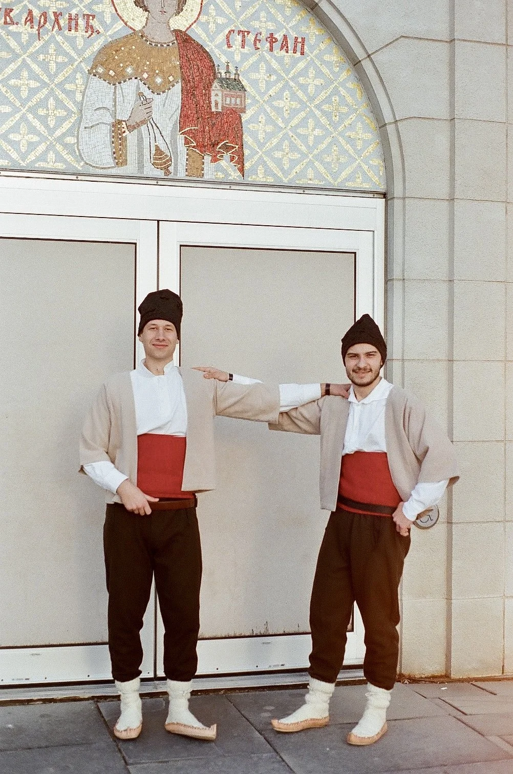 Two young men wearing traditional Albanian clothing, including black hats, white shirts, beige jackets, red sashes, brown pants, and white woolen socks and sandals, stand in front of a wall with a religious mosaic depicting a saint holding a church, surrounded by decorative patterns and Greek letters.