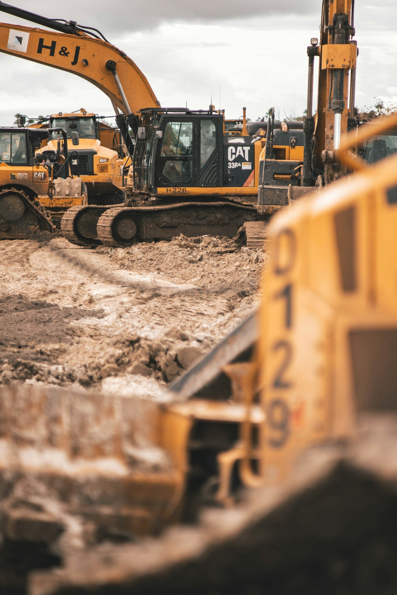 Multiple yellow construction excavators on a muddy construction site under an overcast sky.