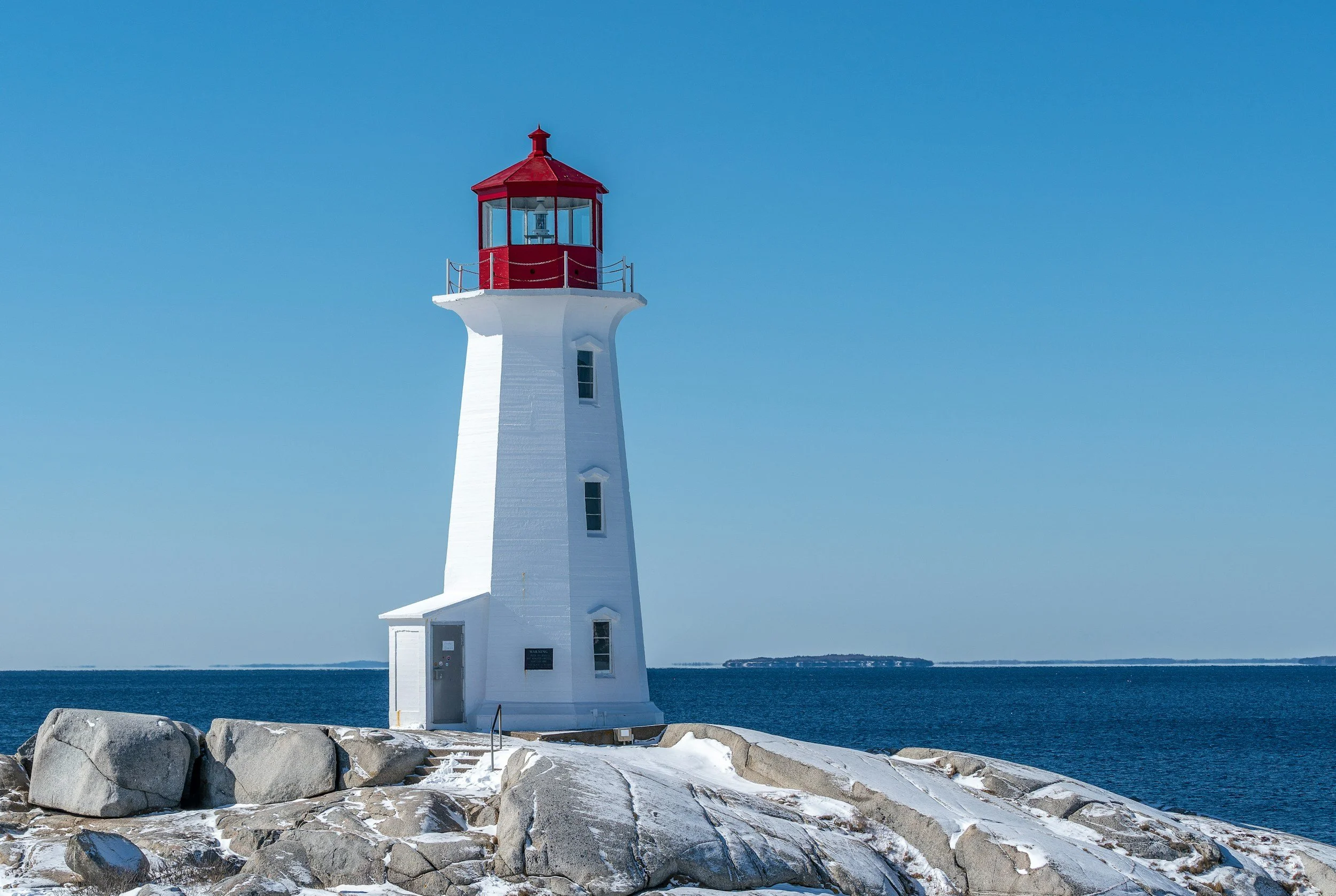 A white lighthouse with a red top and railing, standing on large rocks near the water under a clear blue sky.