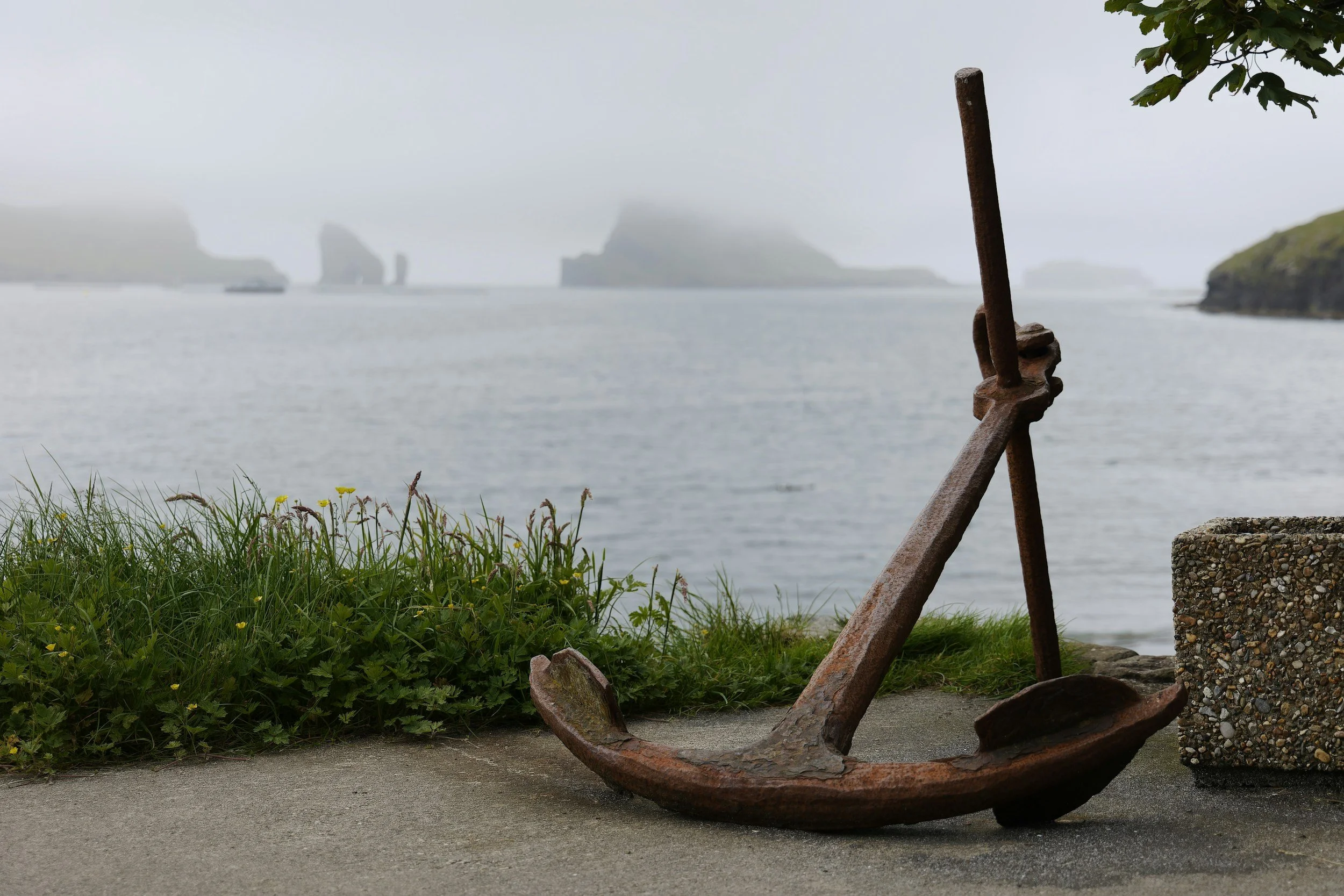 An old rusted anchor lying on the ground near water, with grass, small yellow flowers, rocks, and distant islands shrouded in fog in the background.