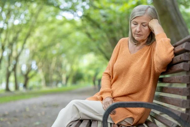 woman with grey hair on a park bench, looking down with a sad look on her face
