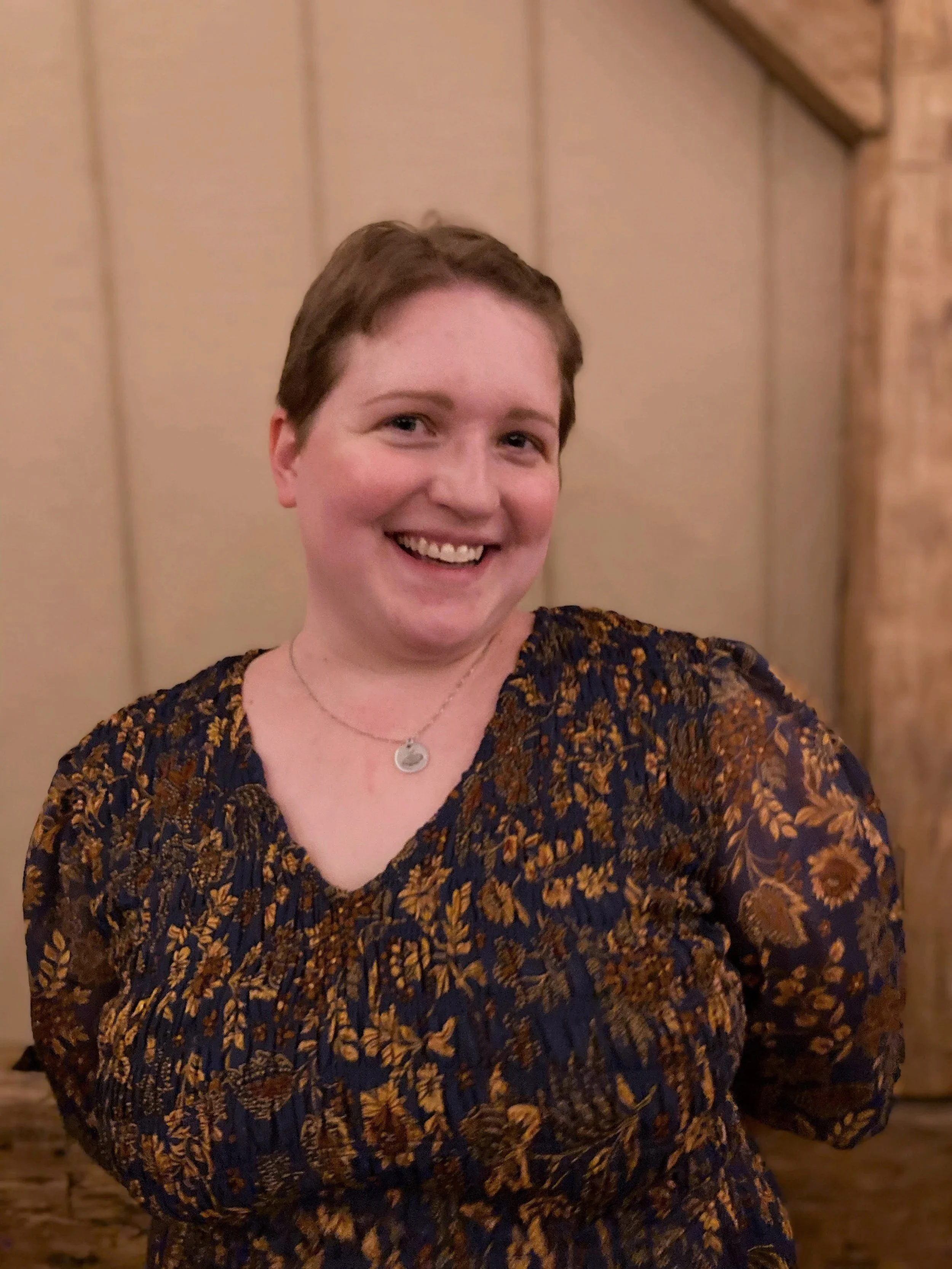 A woman with short brown hair and a fair complexion smiling, wearing a dark floral dress and a silver necklace, standing in front of a beige paneled wall with a wooden trim.