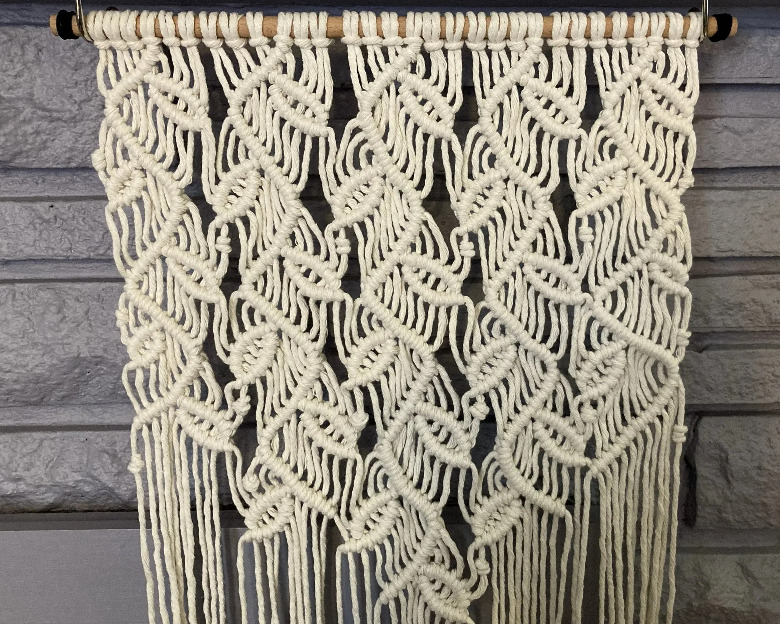Macrame wall hanging with intricate knotted leaf patterns hanging from a wooden rod against a gray stone background.