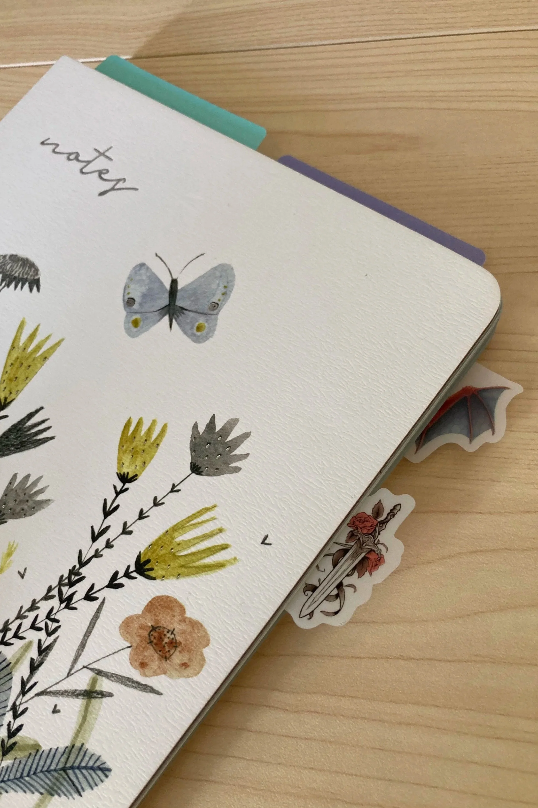 Journal cover with watercolor botanical illustrations and the word 'notes' written at the top, placed on a wooden surface with some bookmarks.
