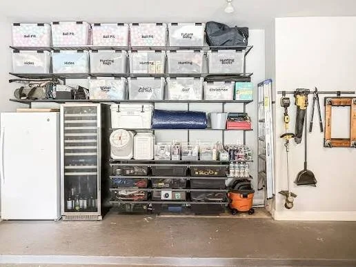 Garage storage with labeled plastic bins on shelves, a white refrigerator, a wine cooler, various tools hanging on the wall, and an orange vacuum on the floor.