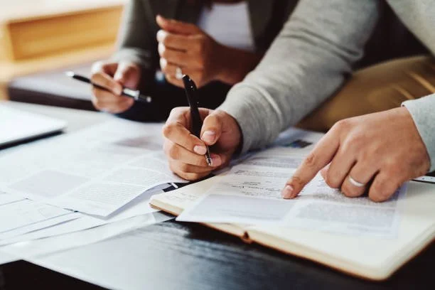 People working at a desk with papers, notebooks, and pens, discussing documents.