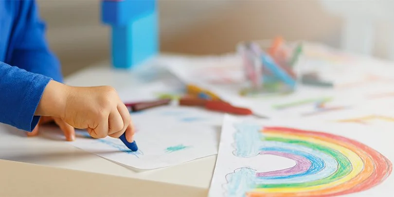 A child's hand drawing on paper with a blue crayon, with colorful rainbow and sky drawings on the table.