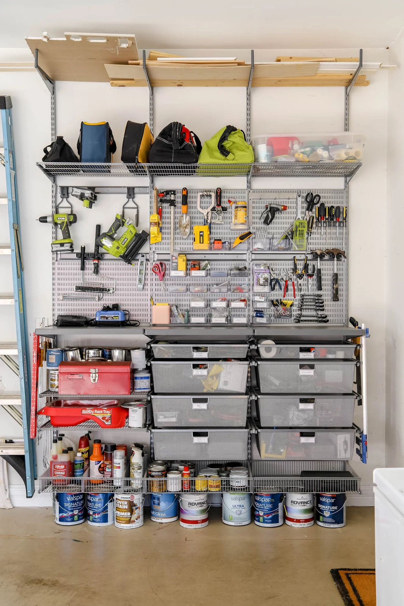 Garage storage wall with organized tools, paints, and supplies. Top shelf has wood planks and bags. Middle shelves contain power tools, hand tools, and storage boxes. The bottom shelf holds paint cans and supplies. Floor is concrete, and a small section of garage floor mat is visible.